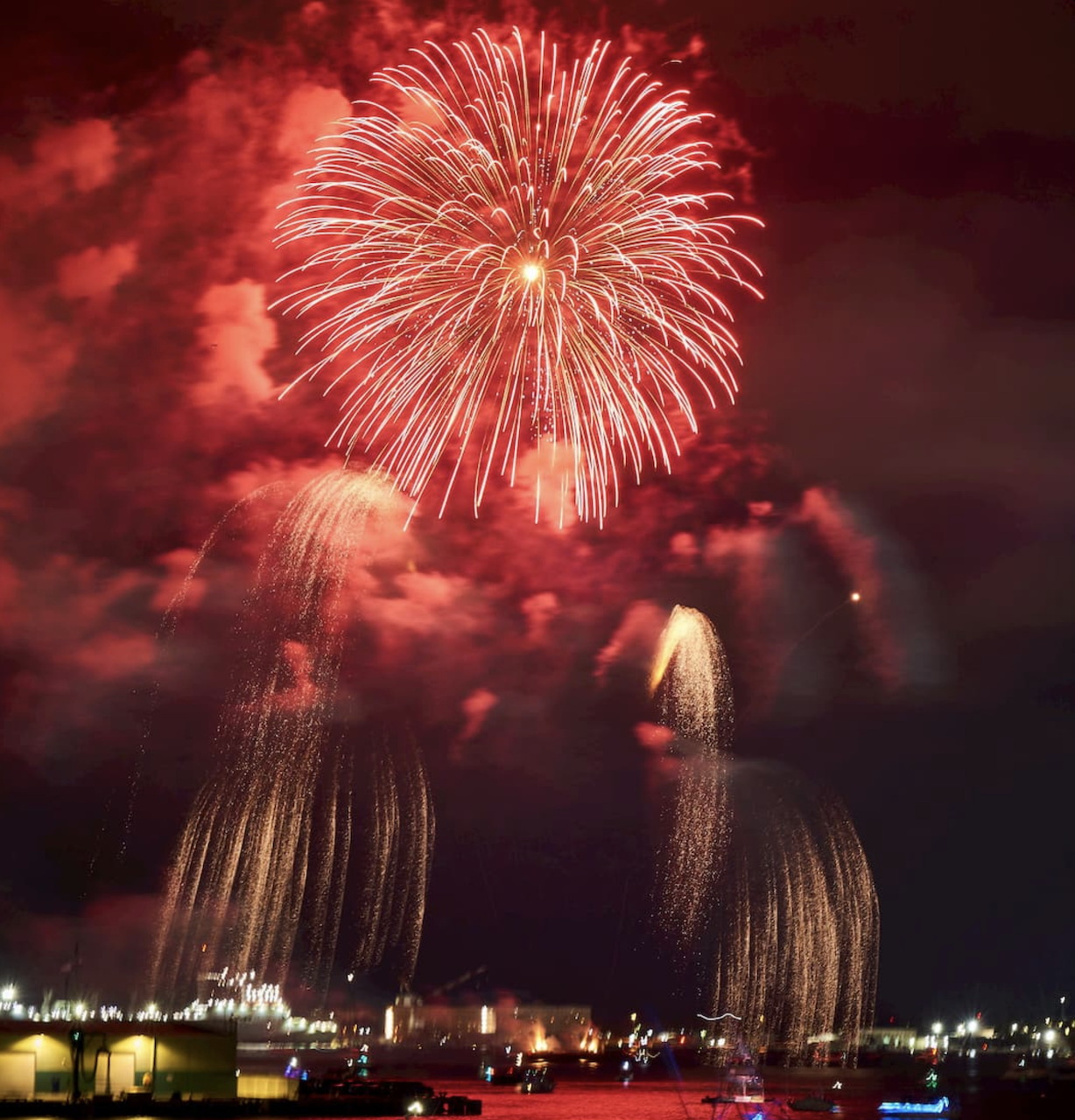 Large red and white fireworks exploding in night sky above waterfront cityscape with water reflections