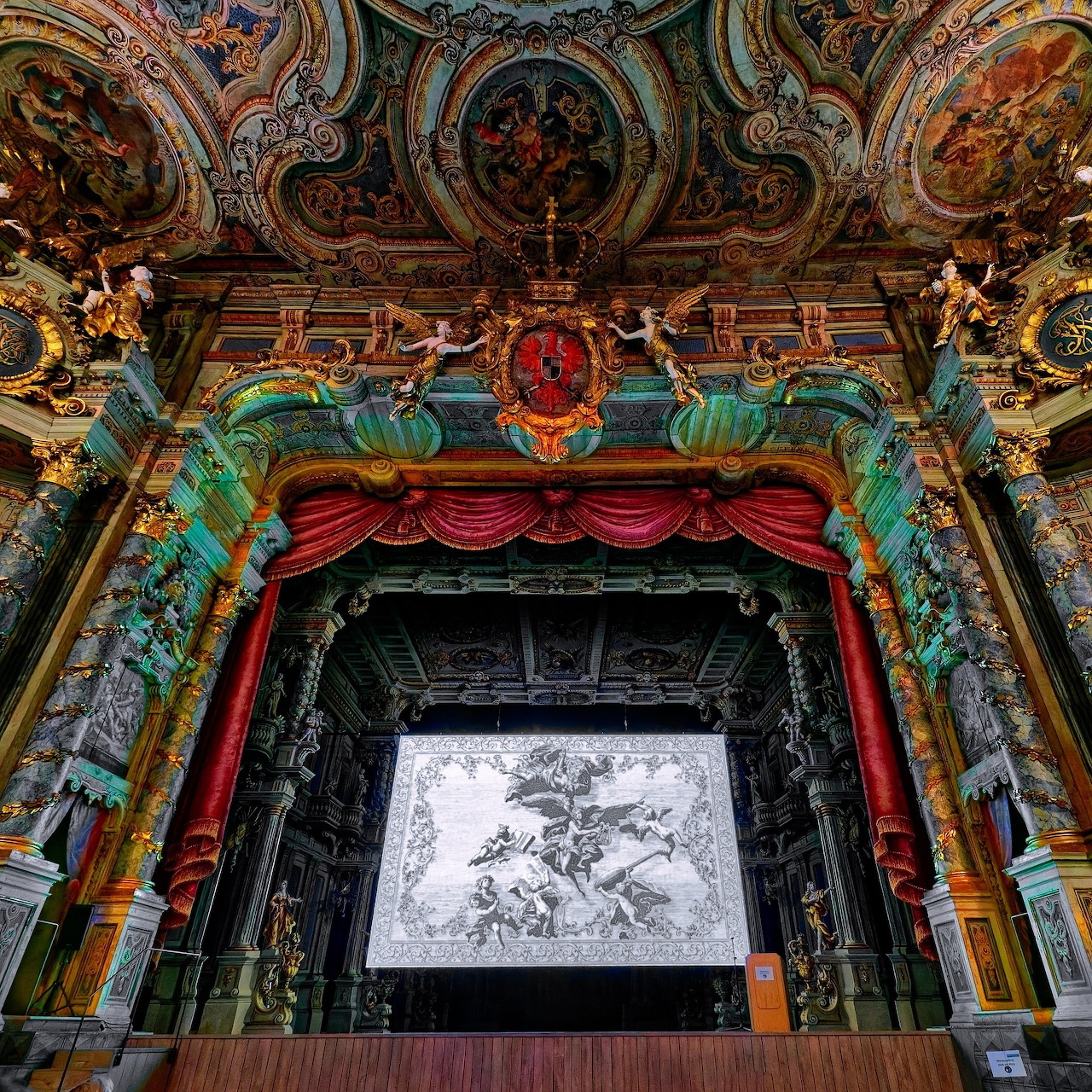 Ornate baroque theater stage with elaborate gilded decorations, painted ceiling, red curtains, and classical architectural details