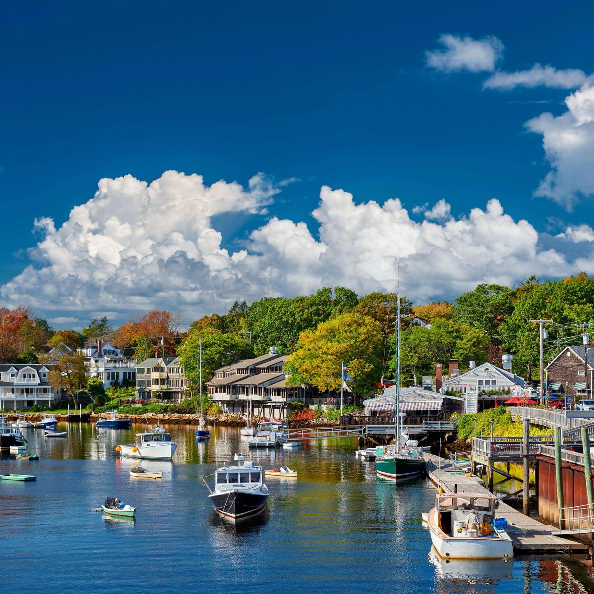 Boats anchored in a scenic harbor with waterfront buildings and green trees under blue sky
