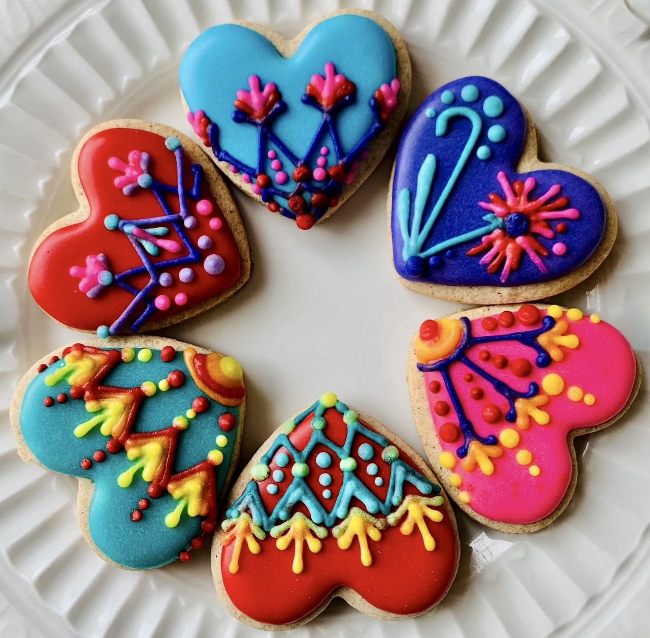 Six decorated heart-shaped cookies with colorful folk art patterns arranged on a white fluted plate