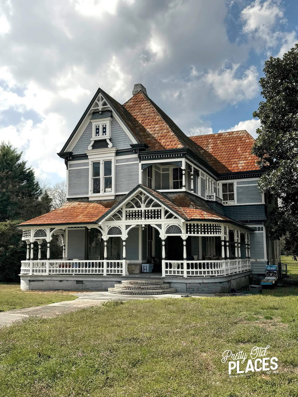 Gray Victorian house with white trim, a wraparound porch, red tile roofs, and a grassy lawn.