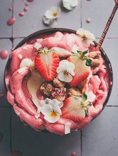 Pink smoothie bowl with strawberry slices, white flowers, coconut, and granola viewed from above