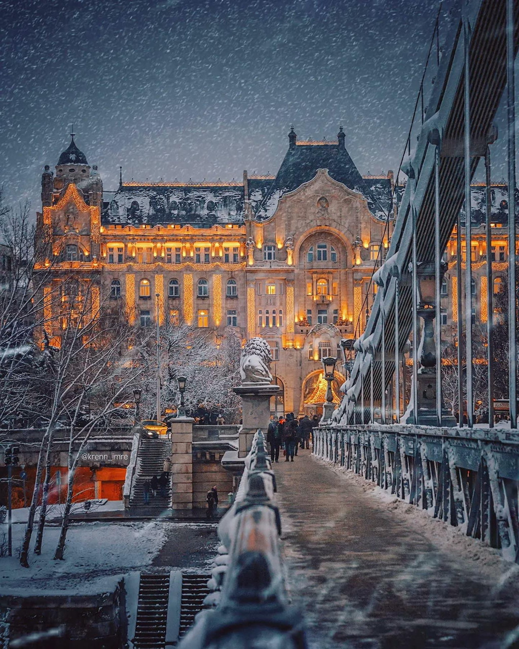 Ornate lit building in Budapest at dusk with snow-covered bridge and pedestrians in winter