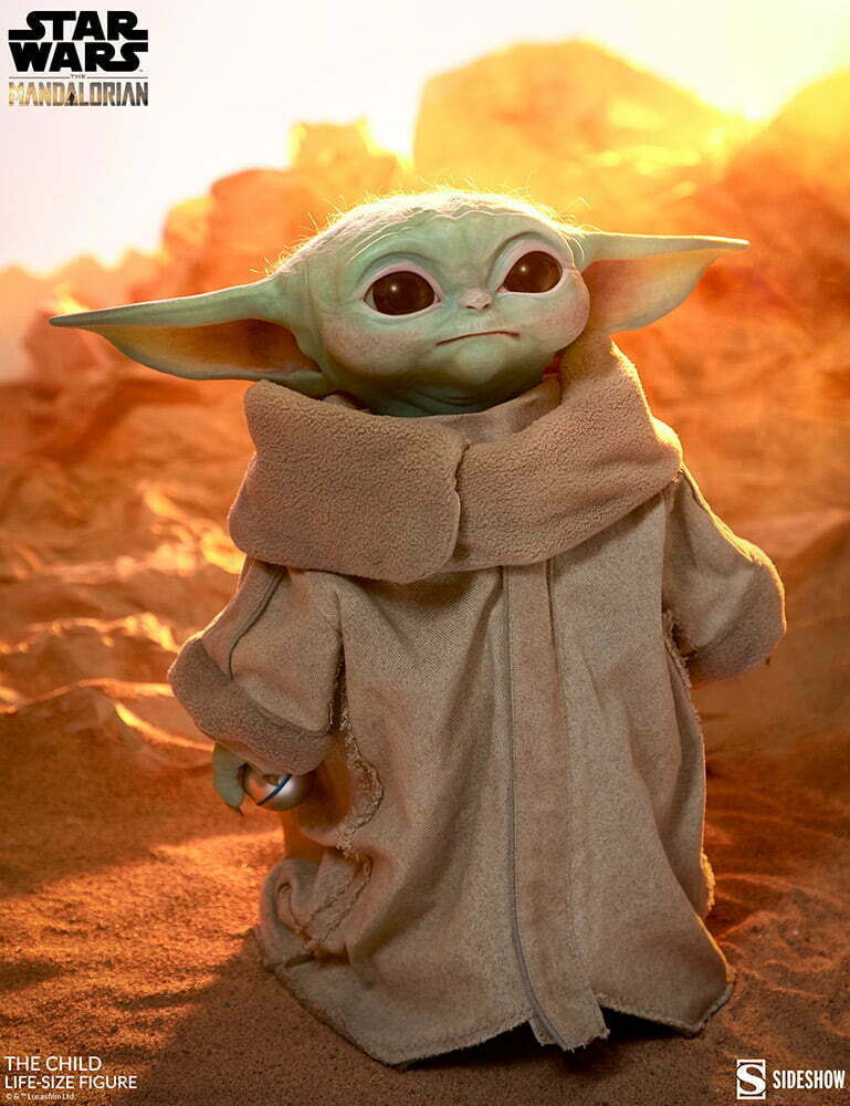 Grogu in a beige robe standing in a sandy desert with a warm orange background