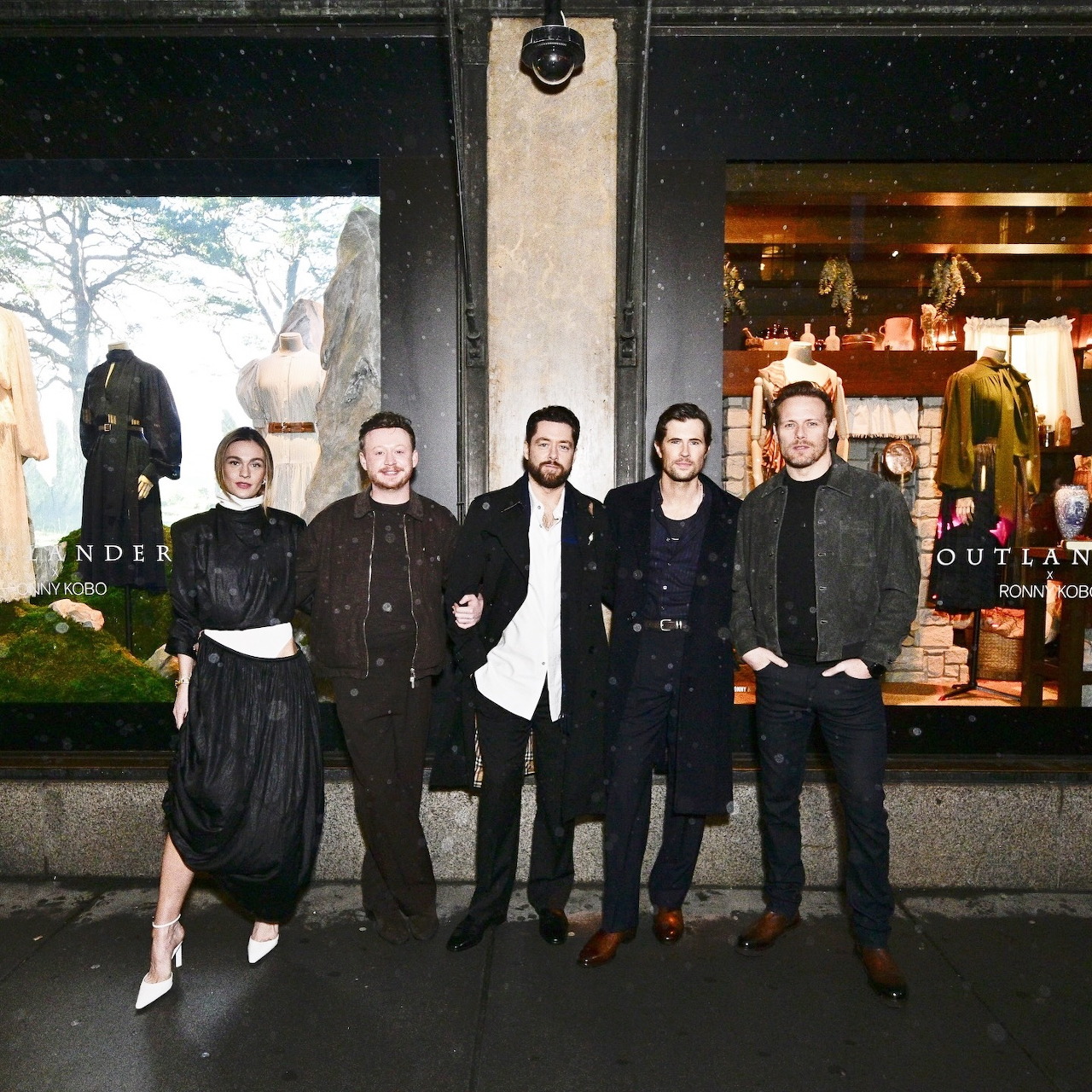Five people posing outside storefront windows with Outlander posters at night