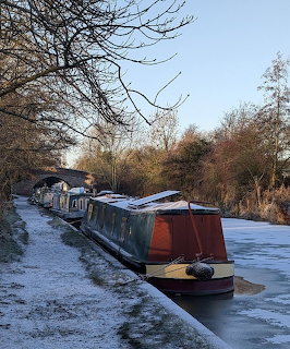 Frozen canal with colorful narrowboats moored along a frosty towpath, stone bridge in background
