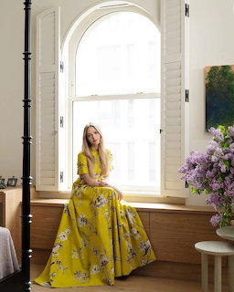 Woman in yellow floral gown seated by tall arched window in bright room with purple flower arrangement
