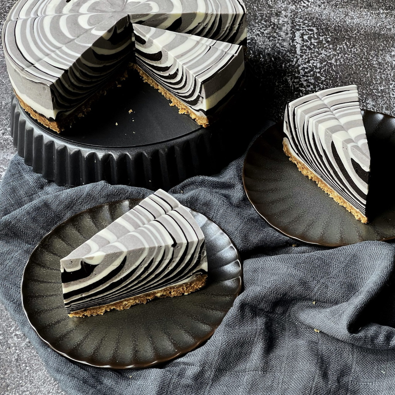 Zebra cheesecake with black and white swirls, served as whole cake and two slices on dark plates
