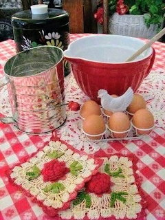 Red mixing bowl with eggs, vintage flour sifter, and crocheted potholders on gingham tablecloth
