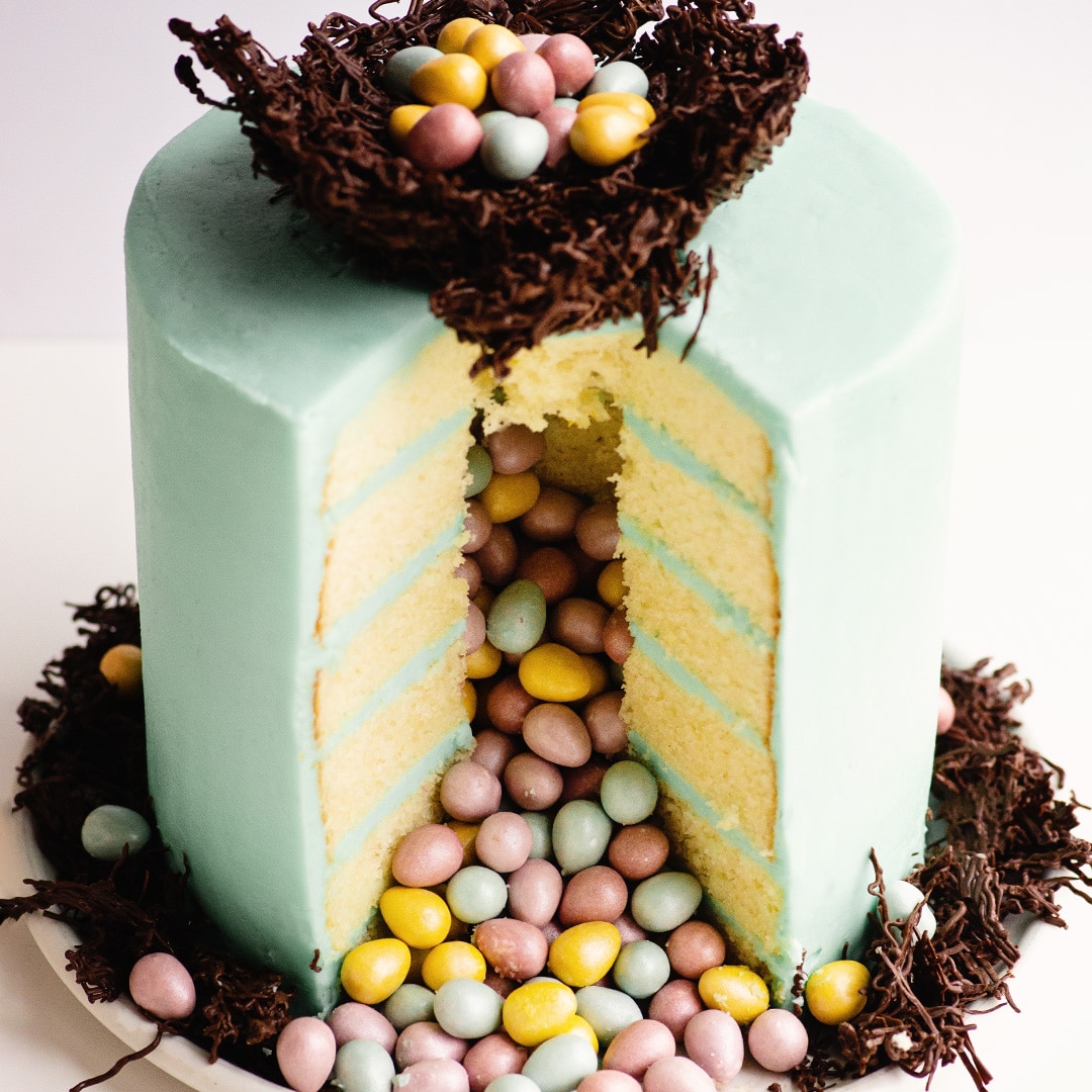 Pastel blue layered cake topped with chocolate nest filled with candy eggs, cut open to reveal candy spilling out