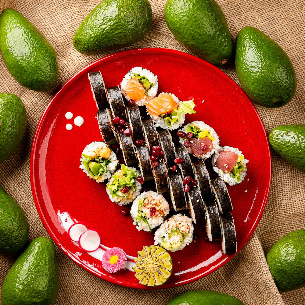 Sushi roll slices with dark sauce on red plate surrounded by avocados