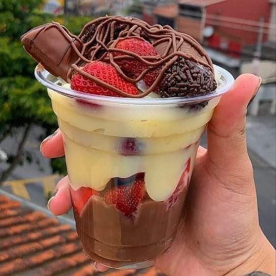 Hand holding layered dessert cup with chocolate, cream, strawberries, and cookie wafers