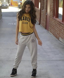 Young woman with curly brown hair in yellow crop top and grey sweatpants posing on a sidewalk