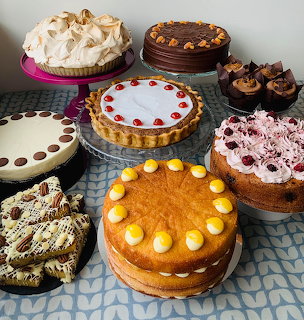 Multiple decorated cakes and pies displayed on checkered blue tablecloth with various toppings and frostings