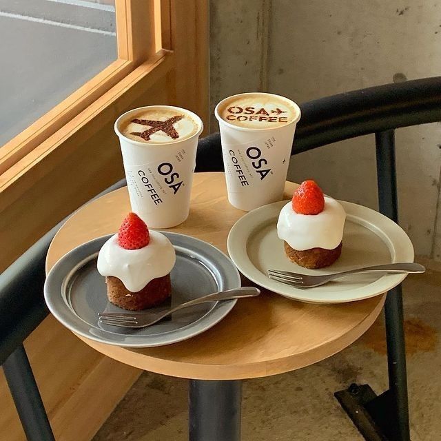 Two coffee cups with OSA branding and two strawberry desserts on gray plates arranged on a round wooden café table