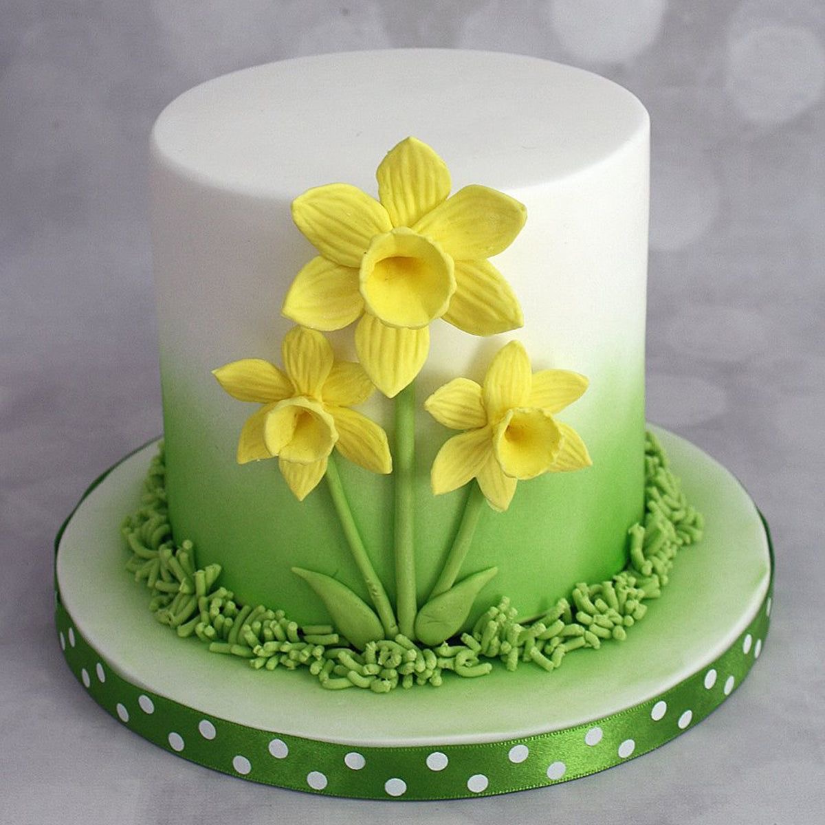Round fondant cake with yellow daffodil decorations on a green ombre base with polka dot ribbon