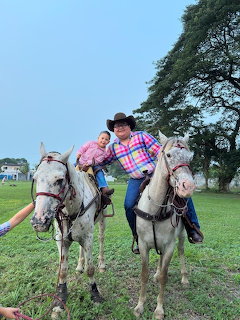Man and child in plaid shirts posing on two white horses on grass under a tree