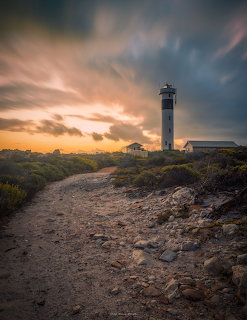 Dirt path leading to a tall white lighthouse under dramatic sunset clouds with coastal vegetation