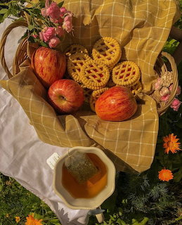 Basket with mini lattice pies, red apples, pink roses, and a cup of tea on yellow gingham cloth outdoors