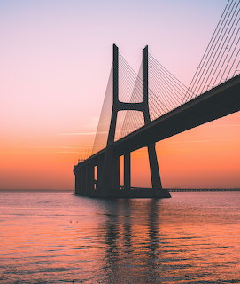 Cable-stayed bridge extending over calm water against a pink and orange sunset sky