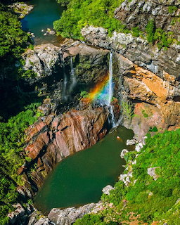 Overhead view of waterfall with rainbow in mist, flowing into turquoise pool surrounded by green cliffs