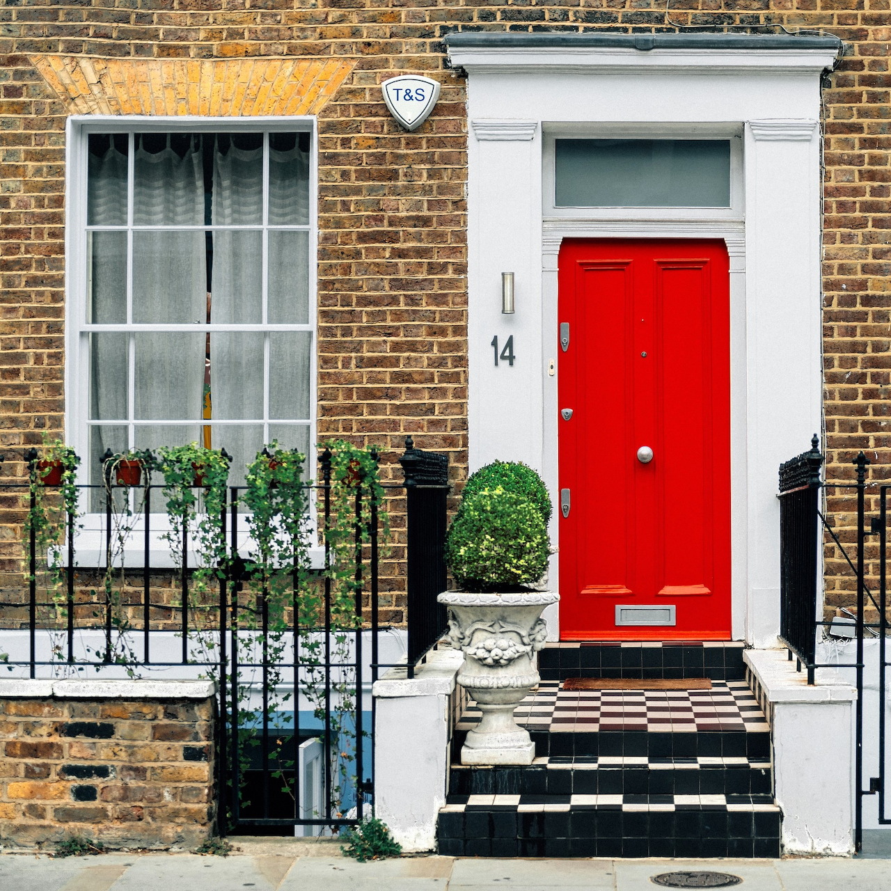 Red door with white trim on brick building facade with window, plants, and checkered steps