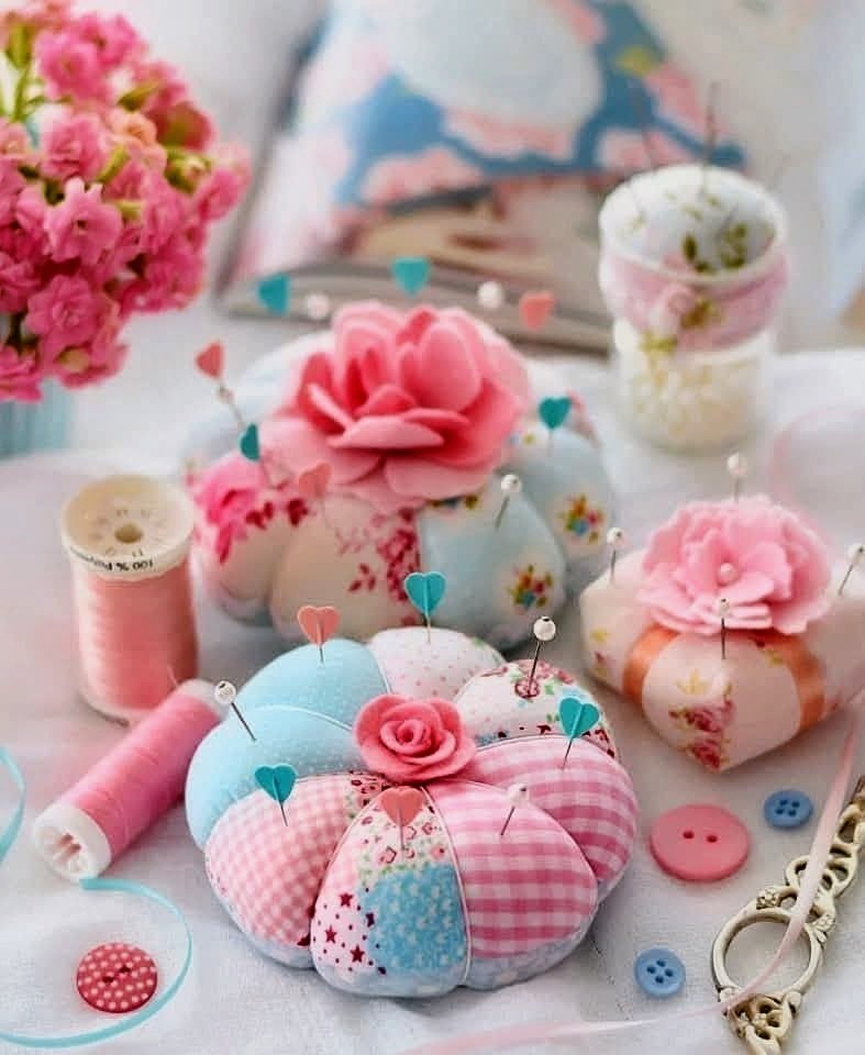 Pastel pink and blue pincushions, thread spools, and buttons arranged on a white surface with floral decorations.