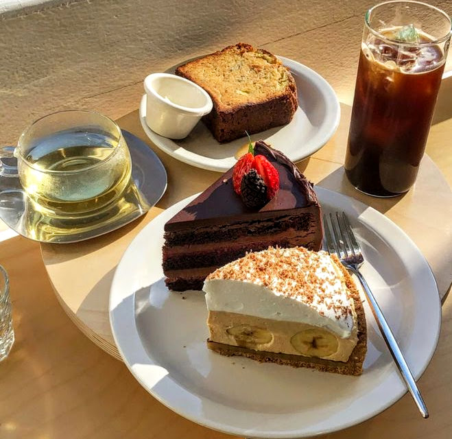 Three white plates with various cake slices, including chocolate and banana cream pie, alongside coffee and tea drinks on a table