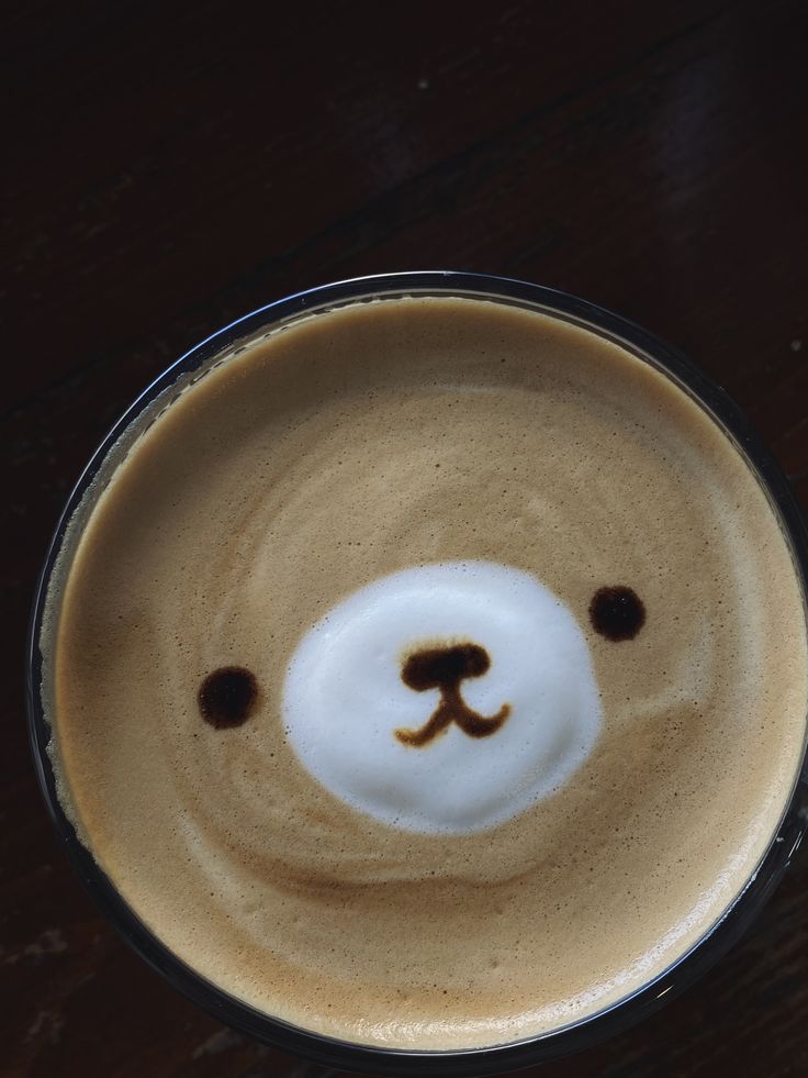 Top-down view of a latte with bear face latte art in a glass cup on a dark wooden table