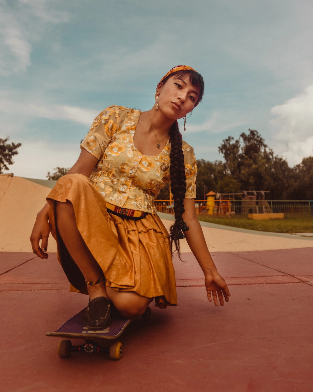 Young Bolivian woman in floral blouse and gold skirt crouching on a skateboard at a skate park