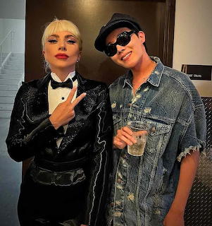 Two people posing together, one in sequined black outfit with bow tie, other in denim jacket with sunglasses and drink