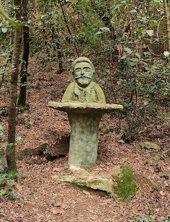 Moss-covered stone bust sculpture of a man on a pedestal in a green forest setting
