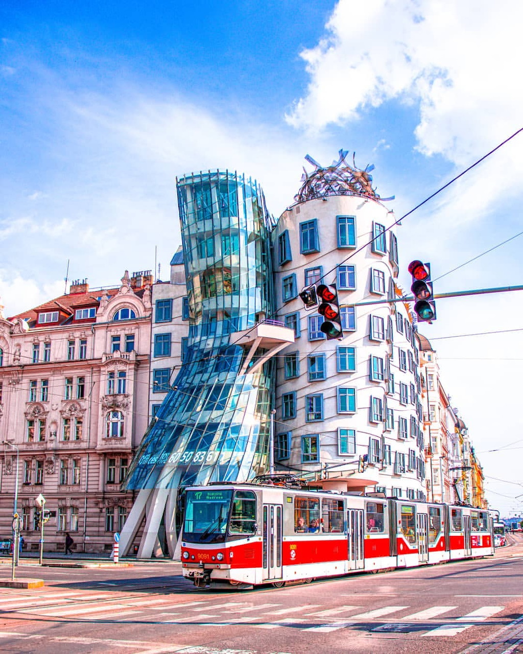 Dancing House building in Prague with curved glass tower and red tram passing in front under blue sky