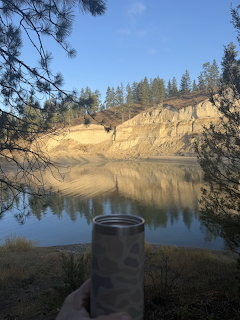 Calm lake reflecting sandy cliffs and pine trees with a hand holding a water bottle in the foreground
