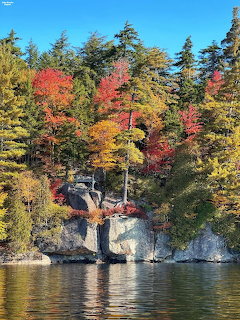 Rocky lakeshore surrounded by colorful autumn trees reflected in calm lake water under blue sky