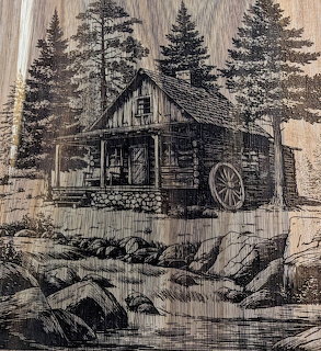 Pen drawing on wood of a log cabin with water wheel among pine trees and a rocky stream