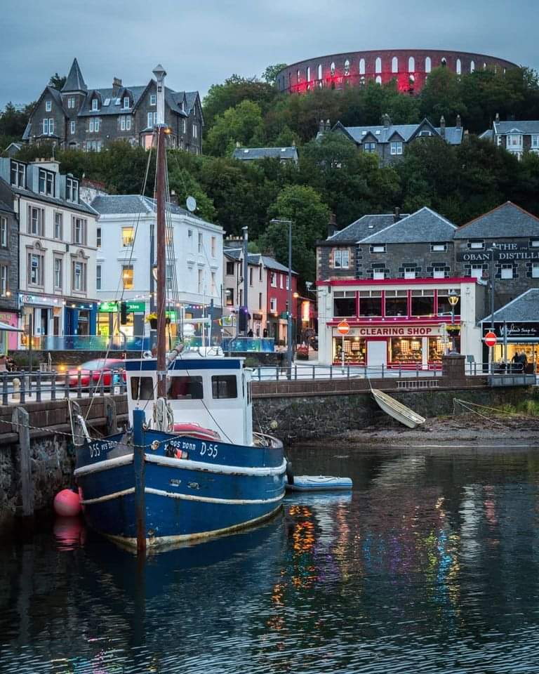 Blue fishing boat in Oban harbour with colorful buildings and McCaig's Tower on hill
