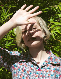 Blonde young woman outdoors with hand raised over her face in bright sunlight and green leaves behind
