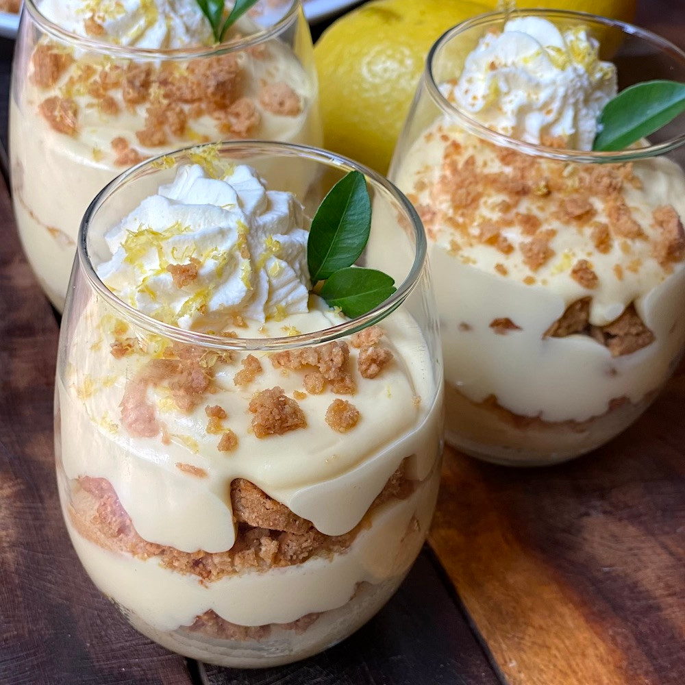 Three layered dessert parfaits in glass jars with cream, cake crumbles, and lemon slices garnished with mint leaves