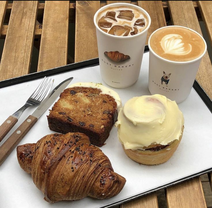 Tray with iced coffee, latte, croissant, banana bread, and a frosted cinnamon roll on a wooden table