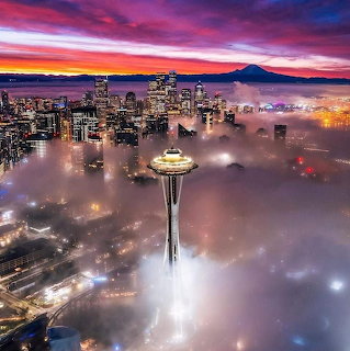 Space Needle tower emerges from fog over Seattle at sunset with mountain backdrop and colorful sky