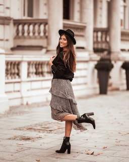 Brunette woman in a black hat and ankle boots posing on a sidewalk by a classic building