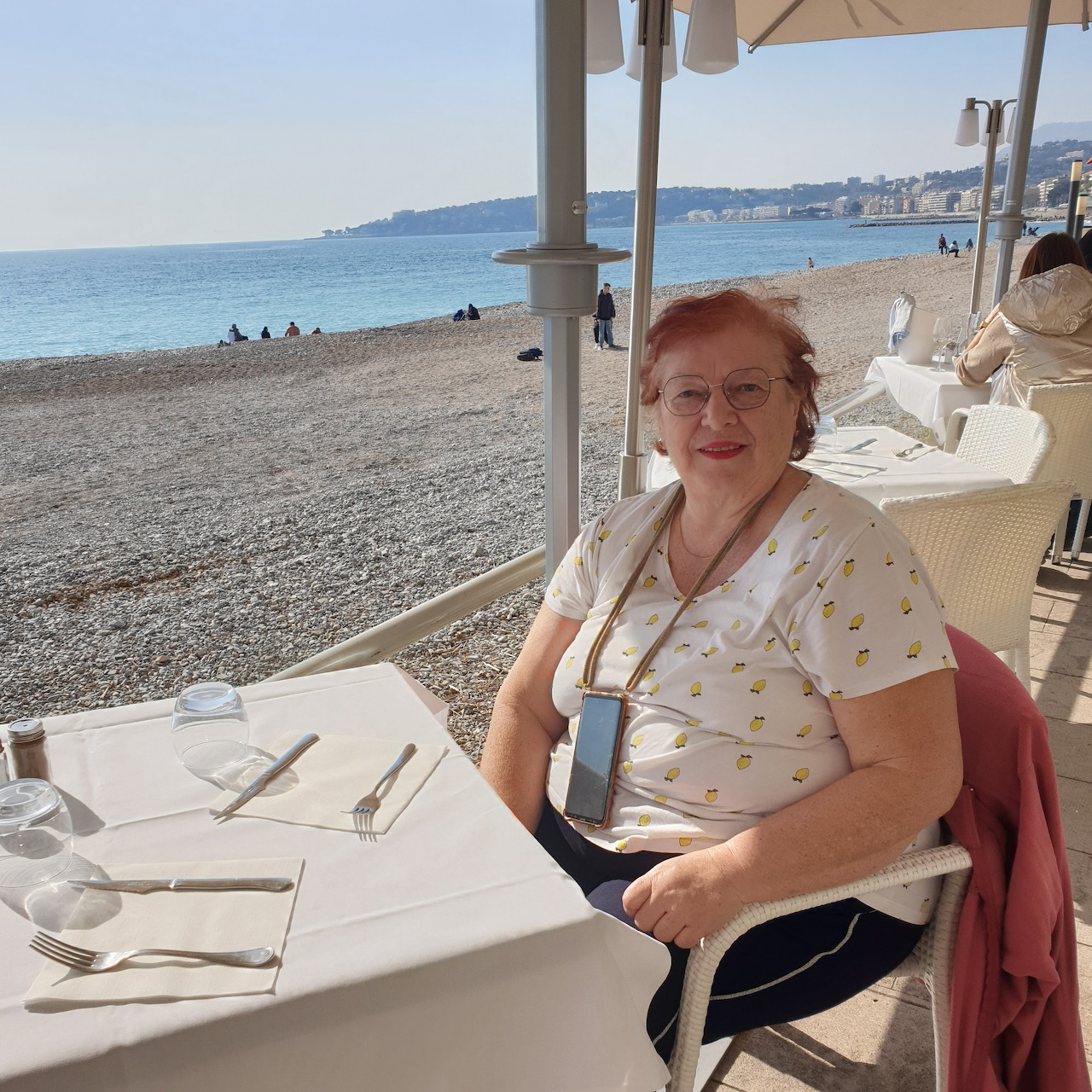 Older woman seated at a beachside cafe table with the sea and pebble beach in the background