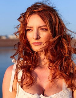 Woman with long red curly hair wearing white top, outdoor portrait in warm sunlight