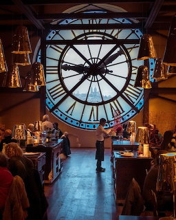 Interior of museum café with large ornate clock window, sculptures, and elegant furnishings in warm light