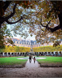 Couple walking through tree-framed square in Paris with historic arcaded buildings and green lawn