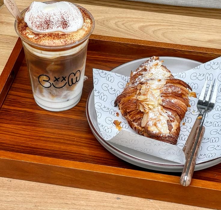 Layered iced coffee with foamy topping next to a powdered croissant on a plate and wooden tray