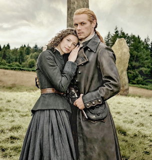 Man and woman in historical Highland dress embracing outdoors near a standing stone