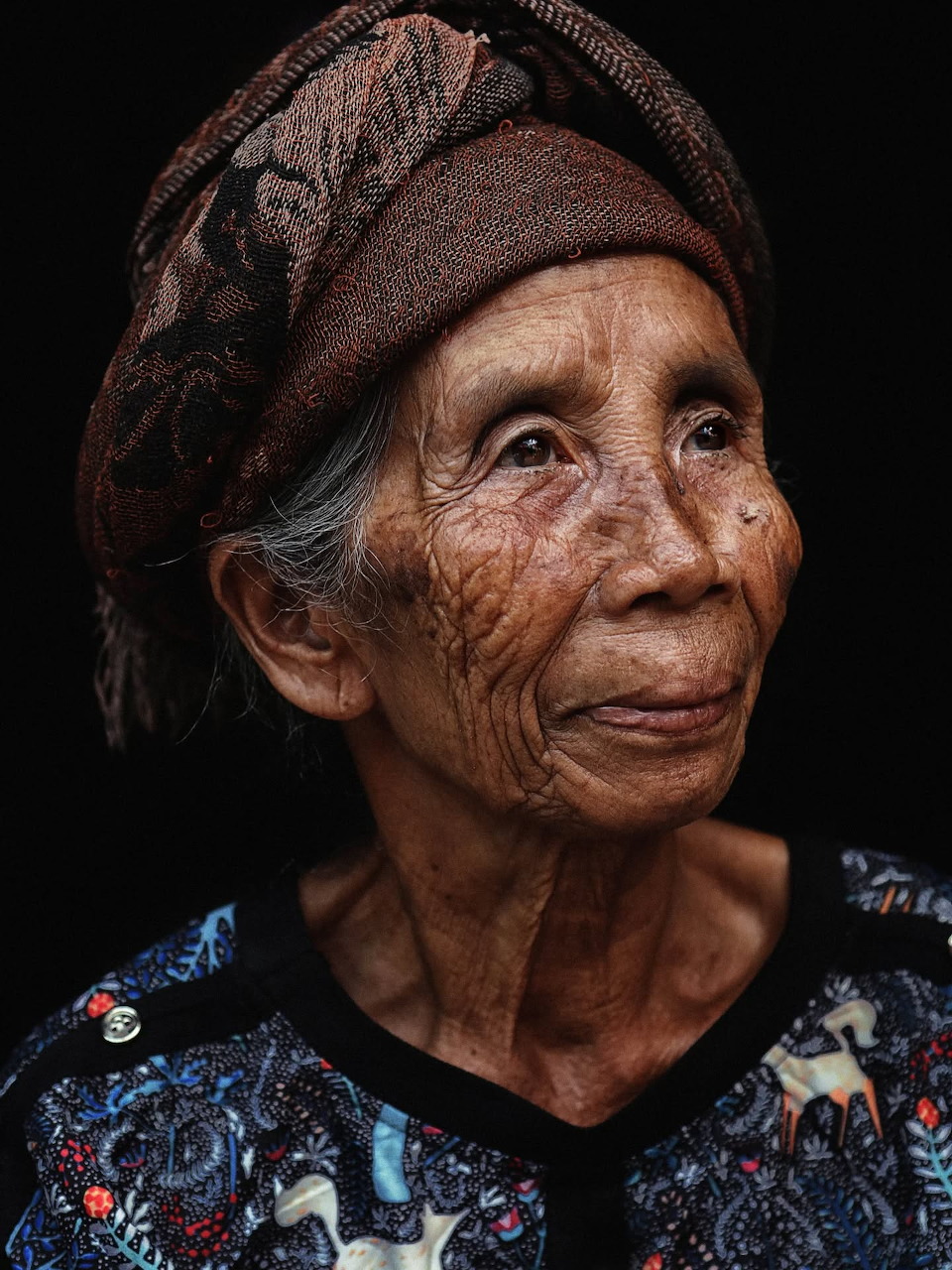 Portrait of an elderly Southeast Asian woman in a patterned headscarf and floral blouse, dark background