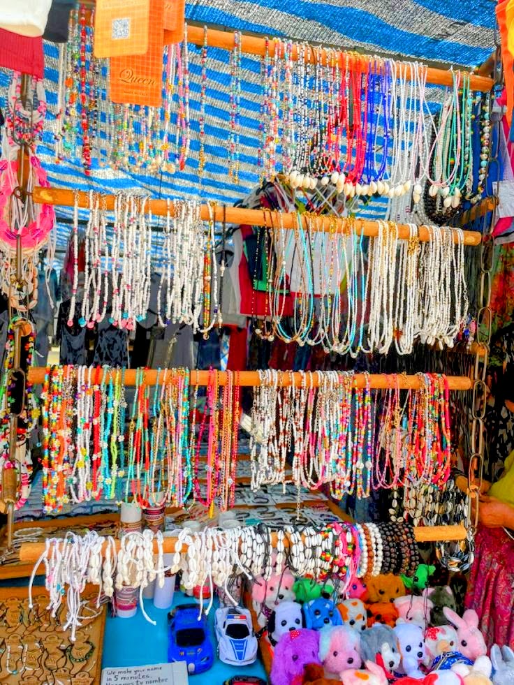 Outdoor market stall with rows of colorful beaded necklaces and small plush toys on display